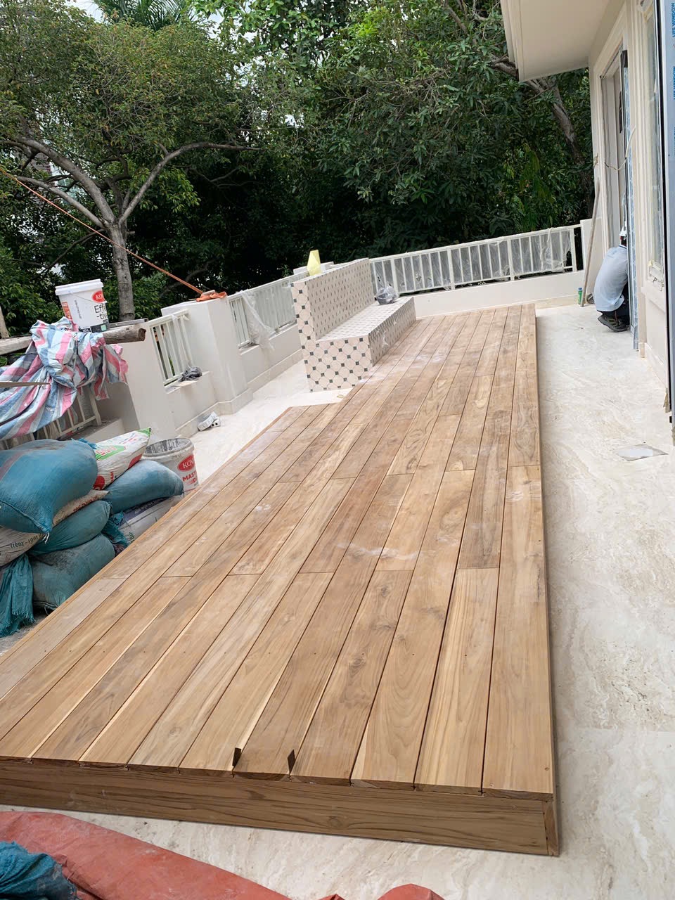 Góc nhìn dọc hiên villa với sàn gỗ bên cạnh cửa nhà, mở về phía cây xanh và hàng rào cuối sân.