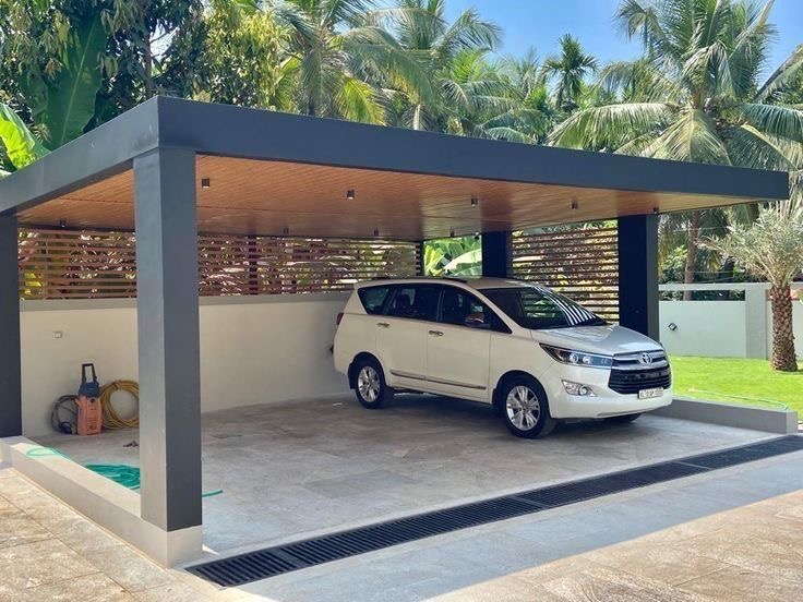 Mái che ô tô trong sân vườn, khung tối màu và trần tông gỗ kín; phía trước có rãnh thoát nước mặt sân.